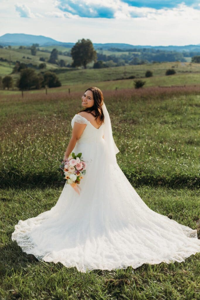 Bride standing with her wedding dress all splayed out on display. She is standing in a sea of green with blue skies and blue rolling mountains in the distance.