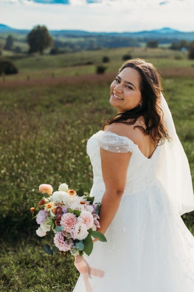 Bride standing with her wedding dress all splayed out on display. She is standing in a sea of green with blue skies and blue rolling mountains in the distance.