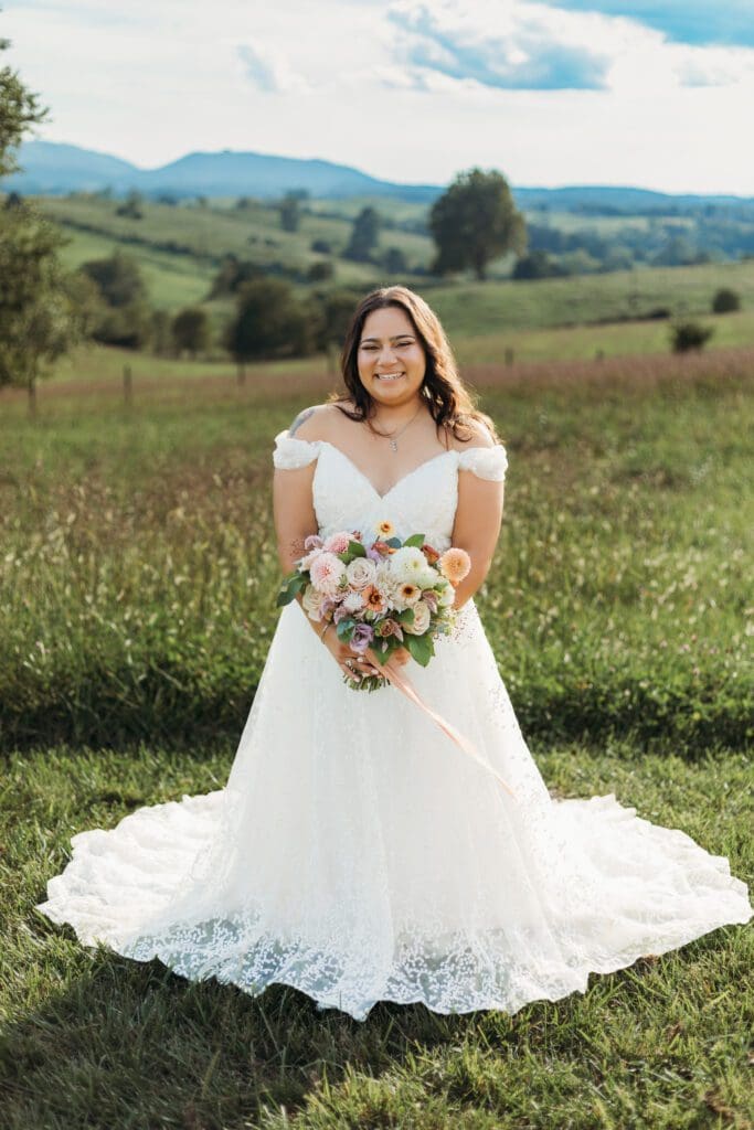 Bride standing with her wedding dress all splayed out on display. She is standing in a sea of green with blue skies and blue rolling mountains in the distance.