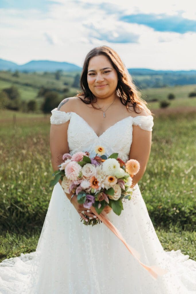 Bride standing with her wedding dress all splayed out on display. She is standing in a sea of green with blue skies and blue rolling mountains in the distance.