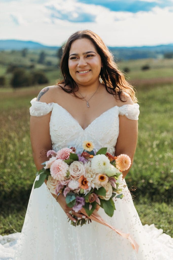 Bride standing with her wedding dress all splayed out on display. She is standing in a sea of green with blue skies and blue rolling mountains in the distance.