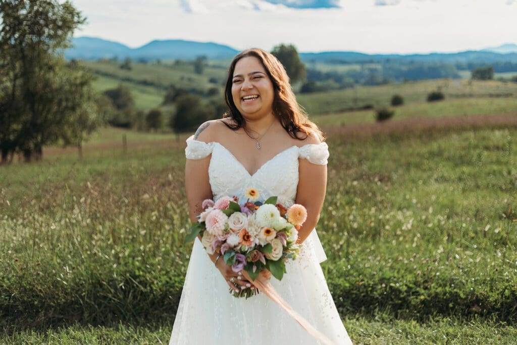 Bride standing with her wedding dress all splayed out on display. She is standing in a sea of green with blue skies and blue rolling mountains in the distance.