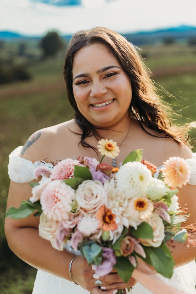 Bride standing with her wedding dress all splayed out on display. She is standing in a sea of green with blue skies and blue rolling mountains in the distance.