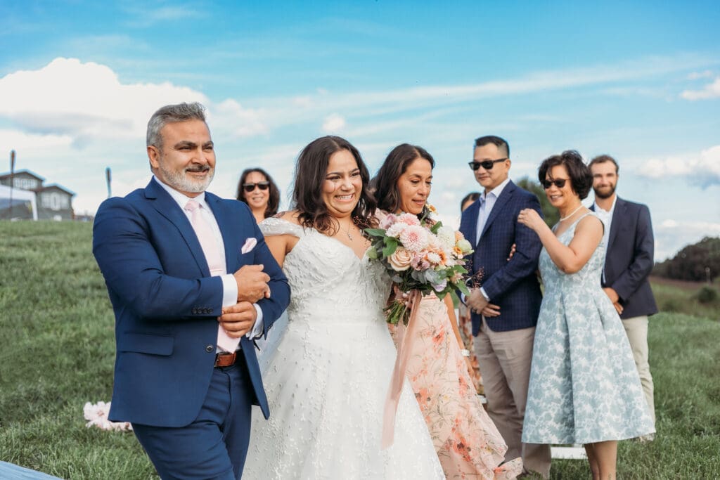 Bride being walked down aisle by her father, surrounded by family with a huge smile on her face.