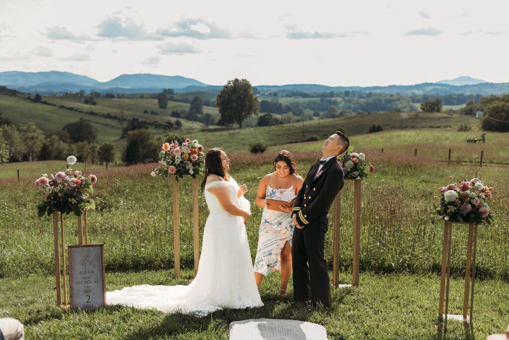 Bride and Groom being married by family member in a field of green with blue rolling mountains in the background. They have a semi circle of flower pedestals around them.