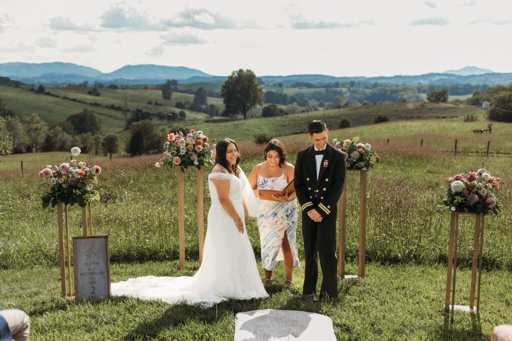 Bride and Groom being married by family member in a field of green with blue rolling mountains in the background. They have a semi circle of flower pedestals around them.