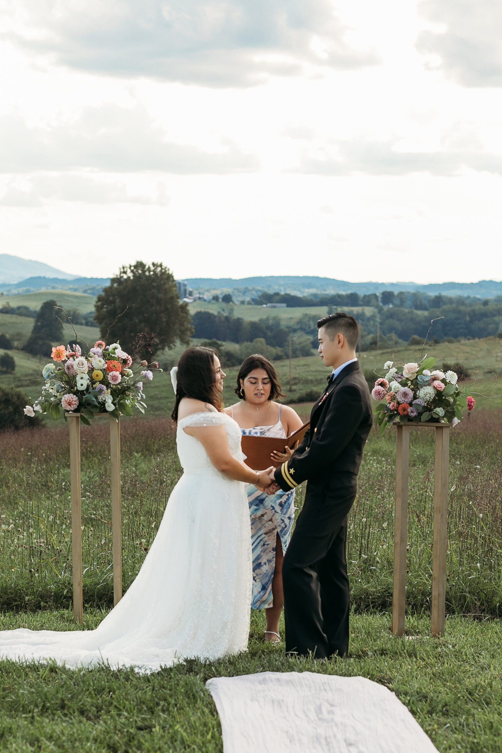 Bride and Groom being married by family member in a field of green with blue rolling mountains in the background. They have a semi circle of flower pedestals around them.