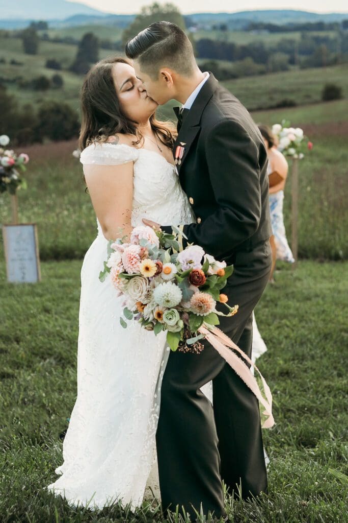 Bride and Groom being married by family member in a field of green with blue rolling mountains in the background. They have a semi circle of flower pedestals around them.