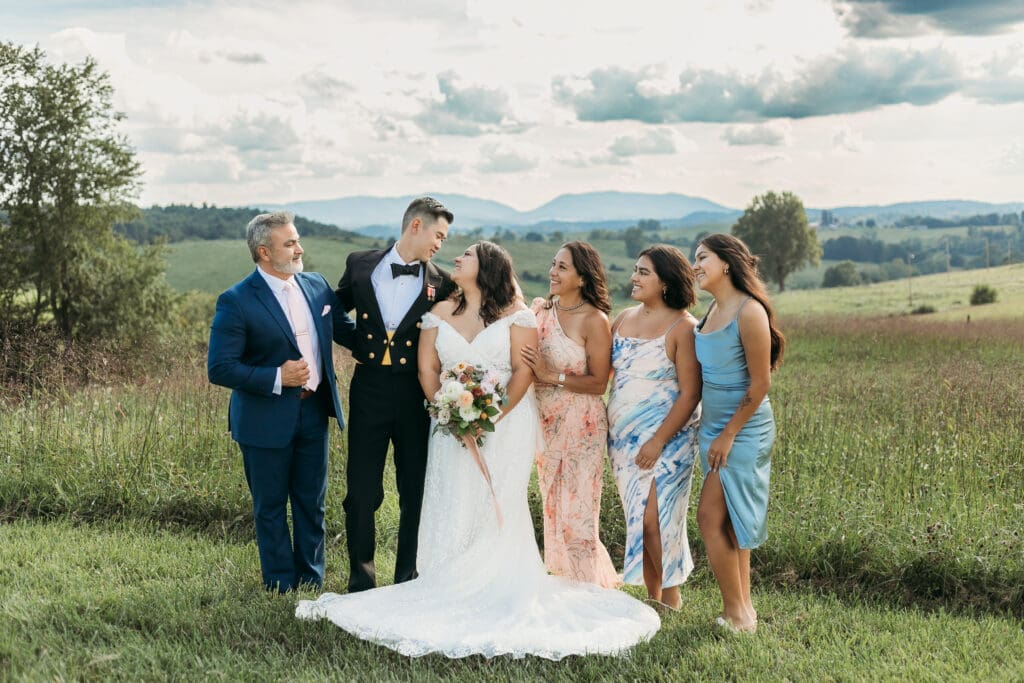 A bride and groom surrounded by family fawning over them in a field of green with rolling green mountains in the background.