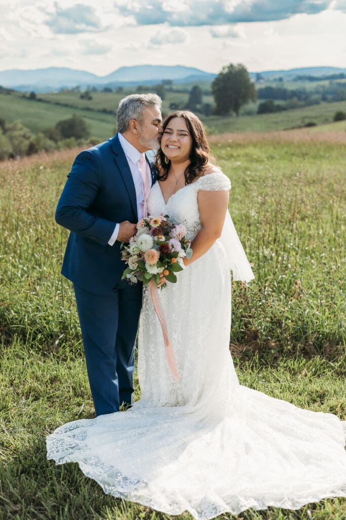 A bride with dad fawning over her in a field of green with rolling green mountains in the background.