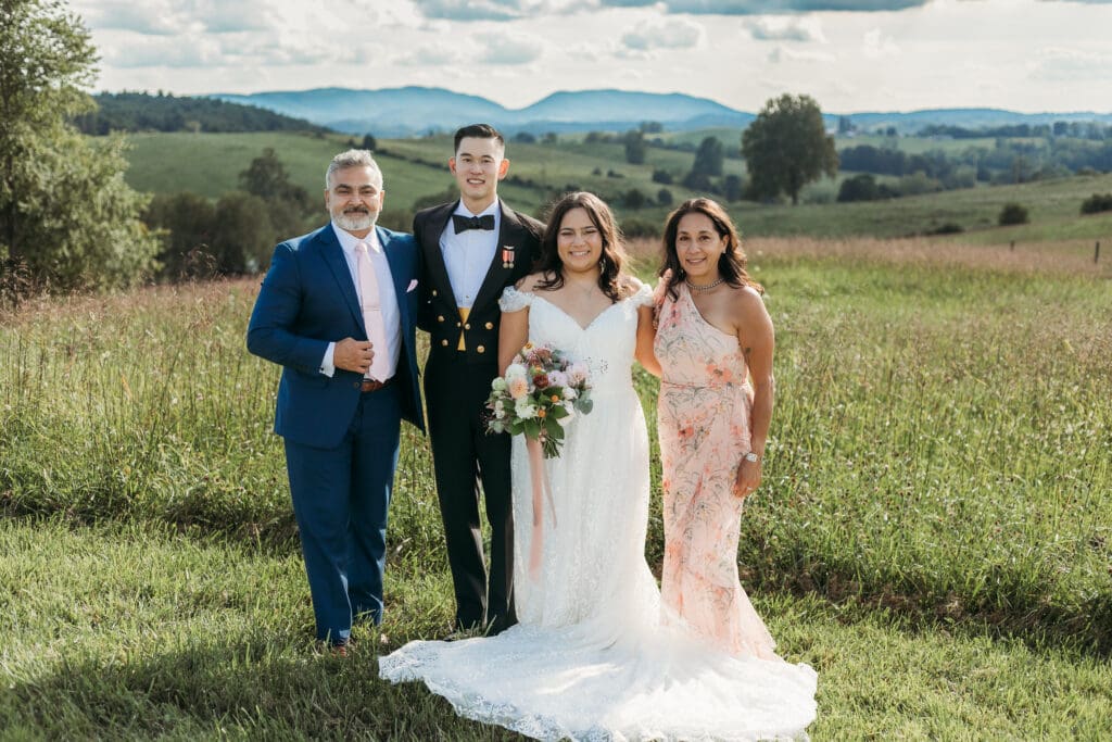 A bride and groom surrounded by family fawning over them in a field of green with rolling green mountains in the background.