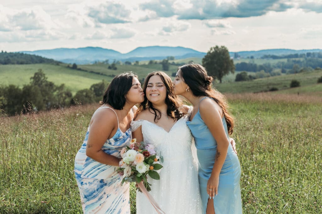 A bride with sisters fawning over her in a field of green with rolling green mountains in the background.