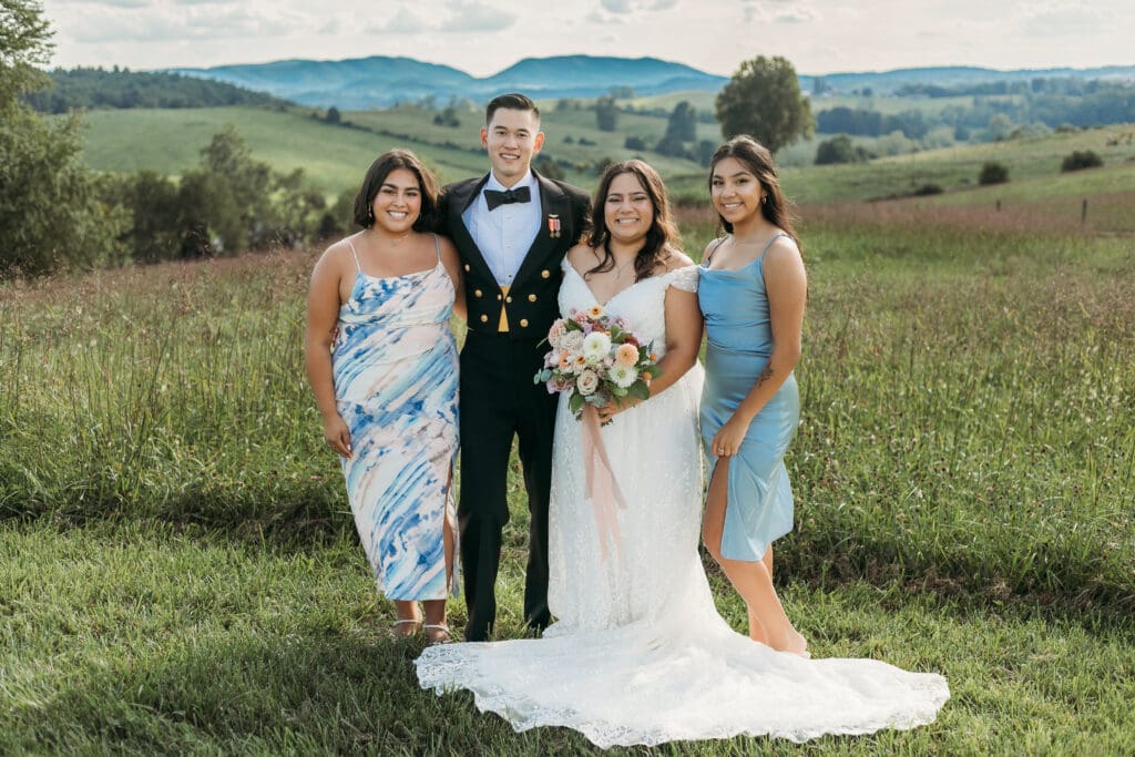 A bride and groom with sisters fawning over her in a field of green with rolling green mountains in the background.