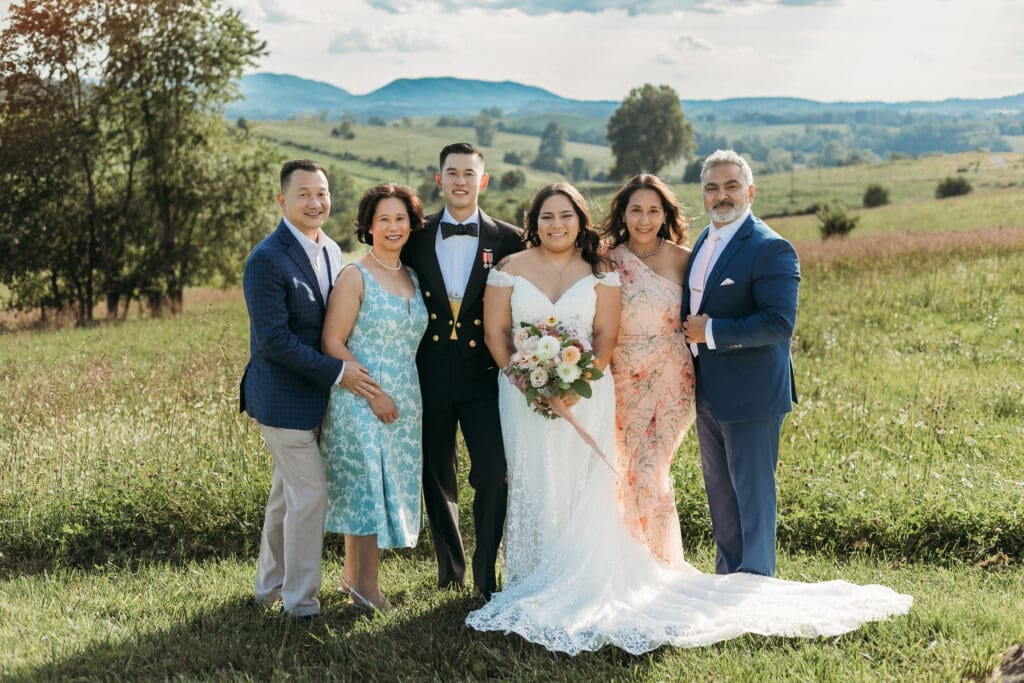 A bride and groom surrounded by family fawning over them in a field of green with rolling green mountains in the background.
