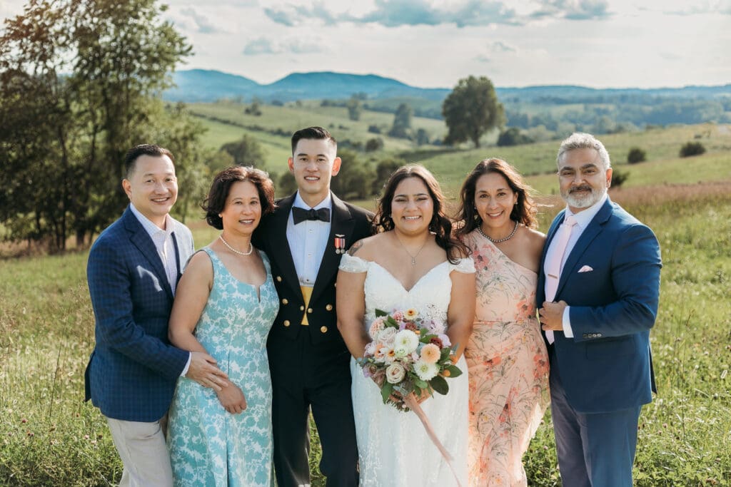 A bride and groom surrounded by family fawning over them in a field of green with rolling green mountains in the background.