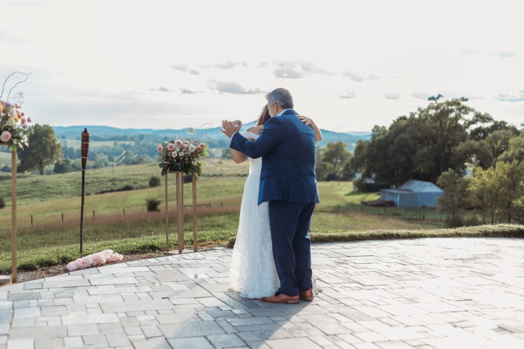Bride having a parent dance with father at mountain elopement. They are dancing on a concrete porch with fields of green surrounding them and rolling blue mountains in the distance.