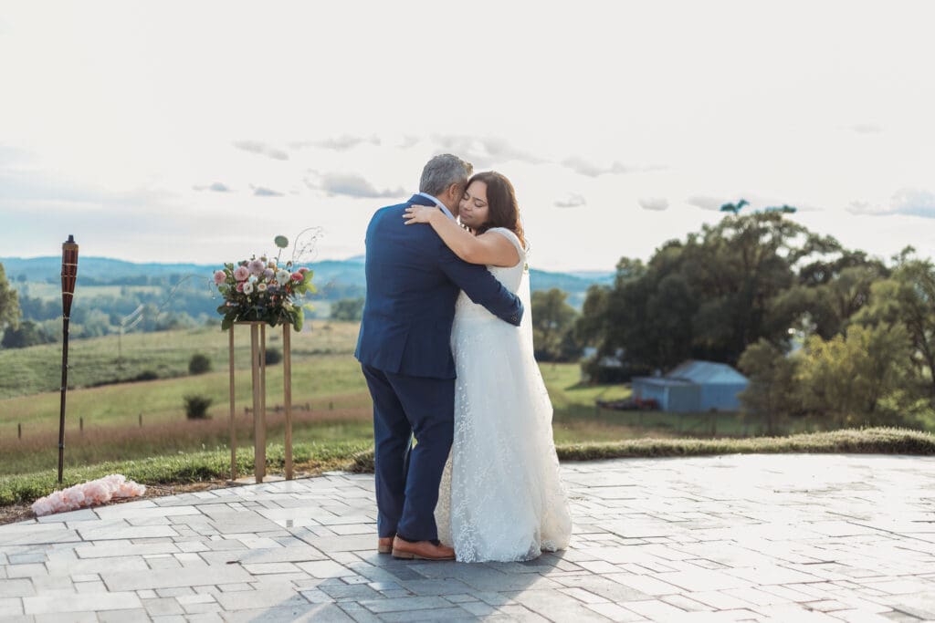 Bride having a parent dance with father at mountain elopement. They are dancing on a concrete porch with fields of green surrounding them and rolling blue mountains in the distance.
