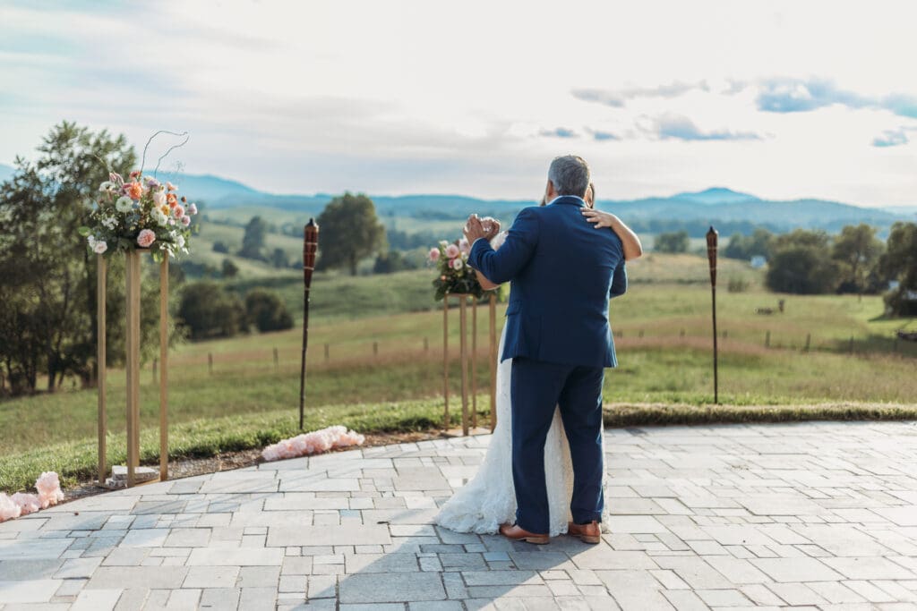 Bride having a parent dance with father at mountain elopement. They are dancing on a concrete porch with fields of green surrounding them and rolling blue mountains in the distance.