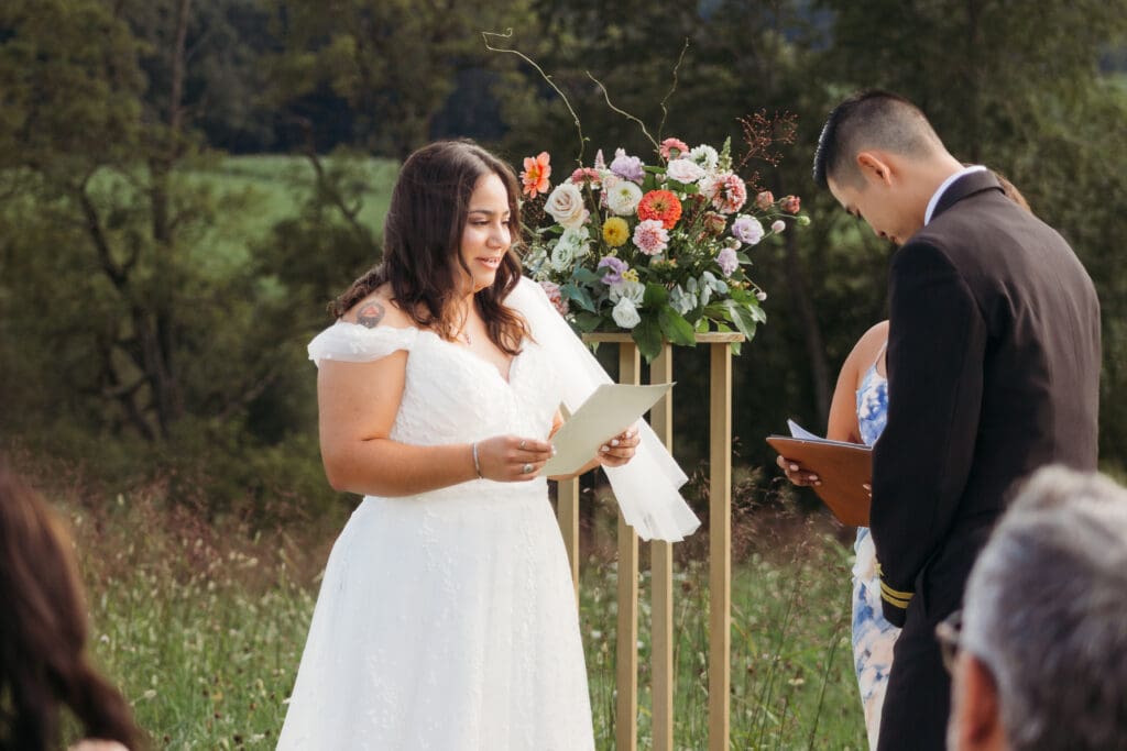 Bride and Groom being married by family member in a field of green with blue rolling mountains in the background. They have a semi circle of flower pedestals around them.