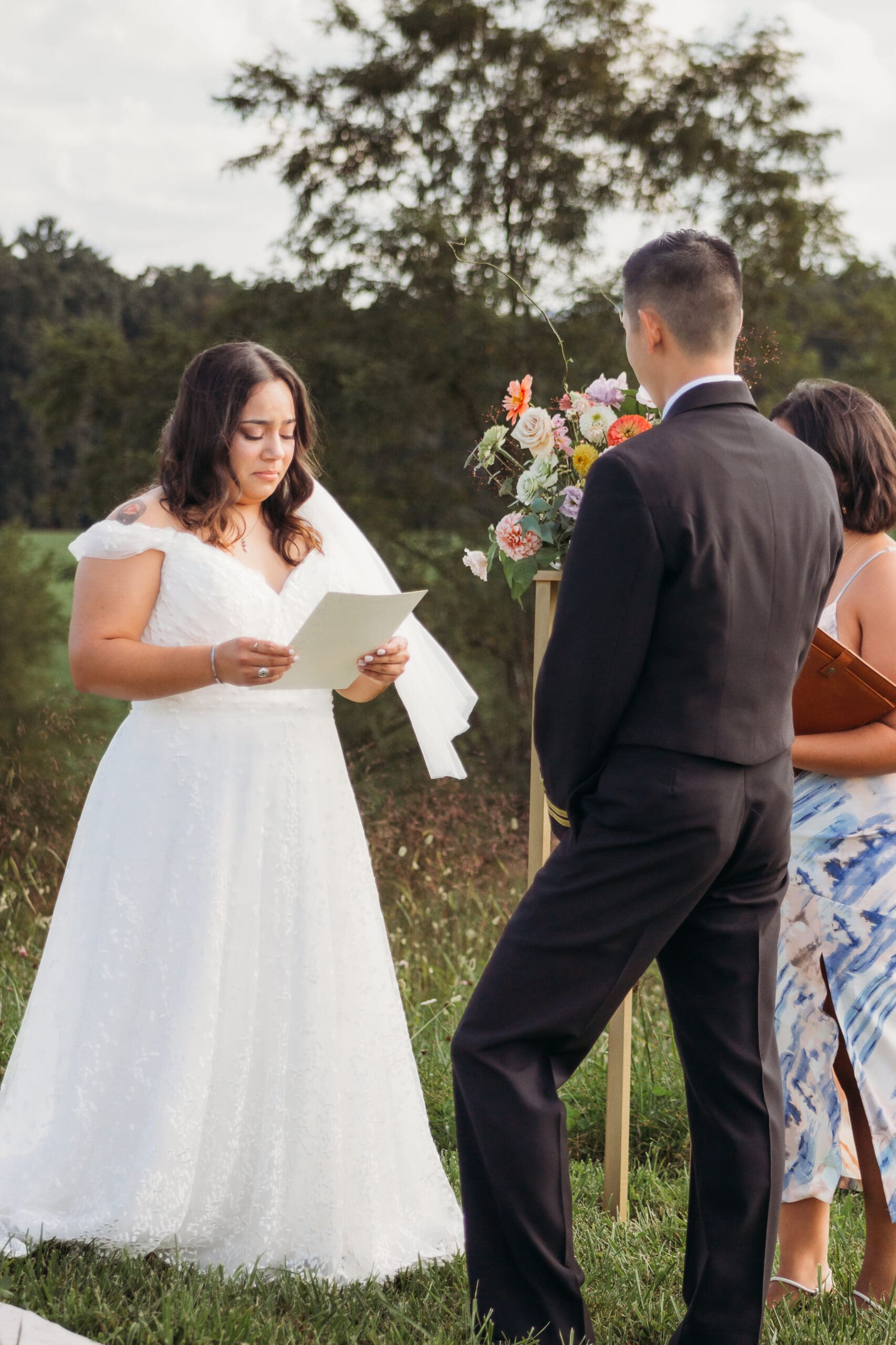 Bride and Groom being married by family member in a field of green with blue rolling mountains in the background. They have a semi circle of flower pedestals around them.
