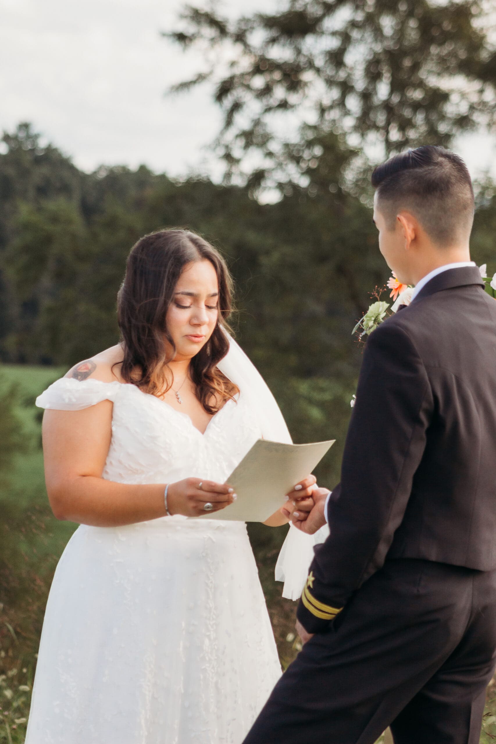 Bride and Groom being married by family member in a field of green with blue rolling mountains in the background. They have a semi circle of flower pedestals around them.