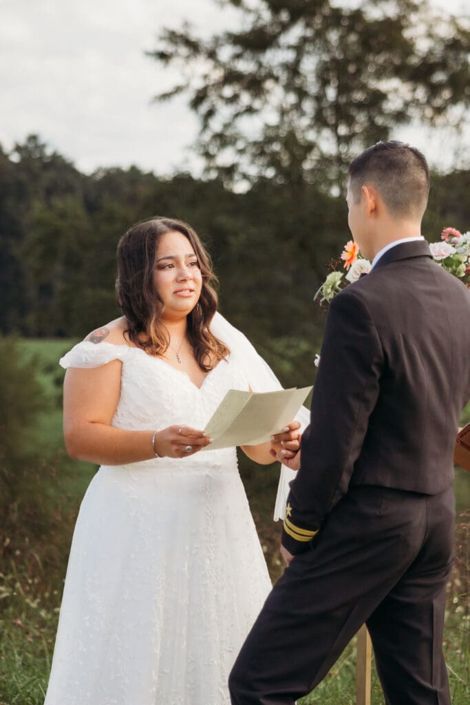 Bride and Groom being married by family member in a field of green with blue rolling mountains in the background. They have a semi circle of flower pedestals around them.