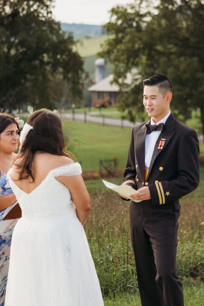 Bride and Groom being married by family member in a field of green with blue rolling mountains in the background. They have a semi circle of flower pedestals around them.