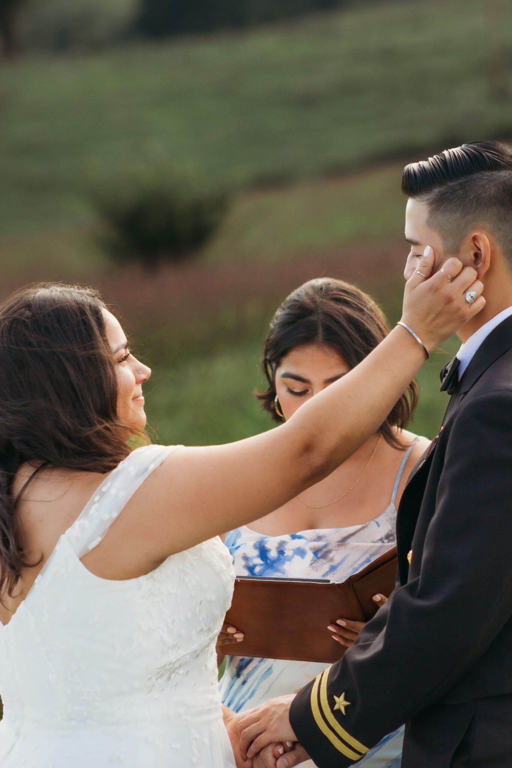 Bride and Groom being married by family member in a field of green with blue rolling mountains in the background. They have a semi circle of flower pedestals around them.