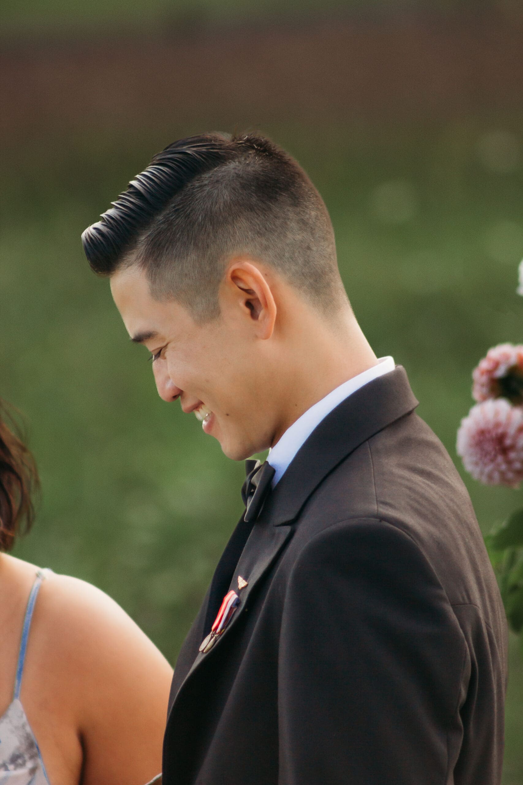 Bride and Groom being married by family member in a field of green with blue rolling mountains in the background. They have a semi circle of flower pedestals around them.