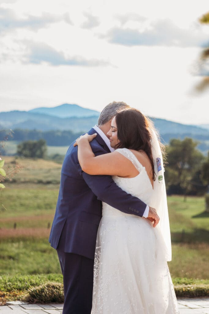 Bride having a parent dance with father at mountain elopement. They are dancing on a concrete porch with fields of green surrounding them and rolling blue mountains in the distance.