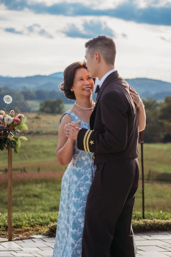 Groom having a parent dance with mother at mountain elopement. They are dancing on a concrete porch with fields of green surrounding them and rolling blue mountains in the distance.