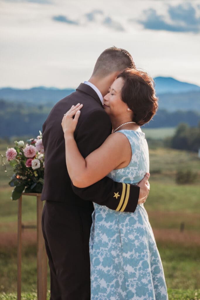Groom having a parent dance with mother at mountain elopement. They are dancing on a concrete porch with fields of green surrounding them and rolling blue mountains in the distance.